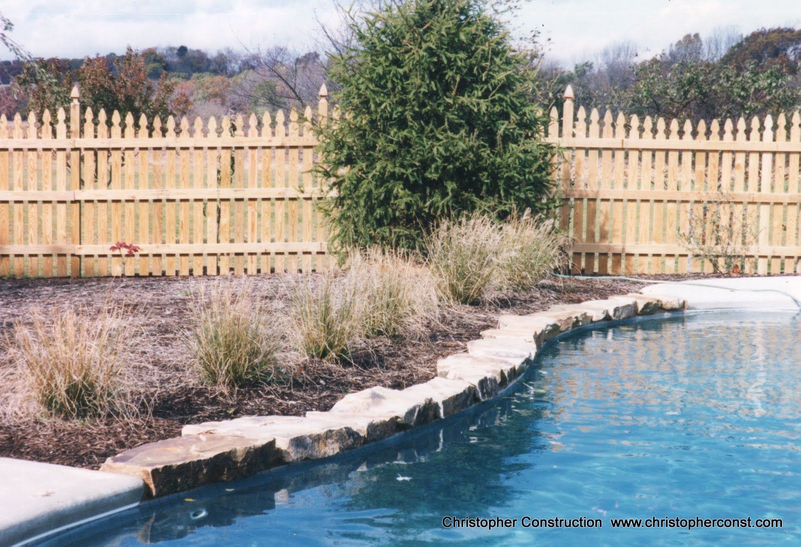 Backyard pool with blue water, stone edge, shrubs, and a wooden picket fence