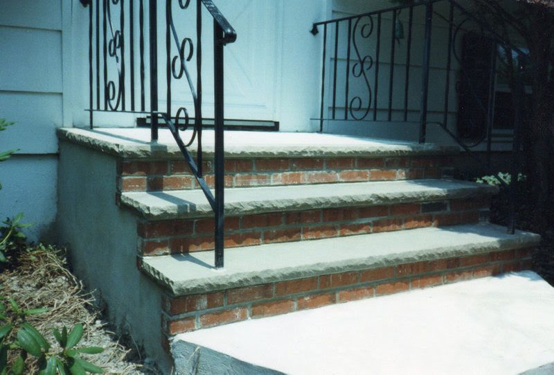Front steps with red brick treads, concrete sides, and black wrought-iron railings leading to a porch.