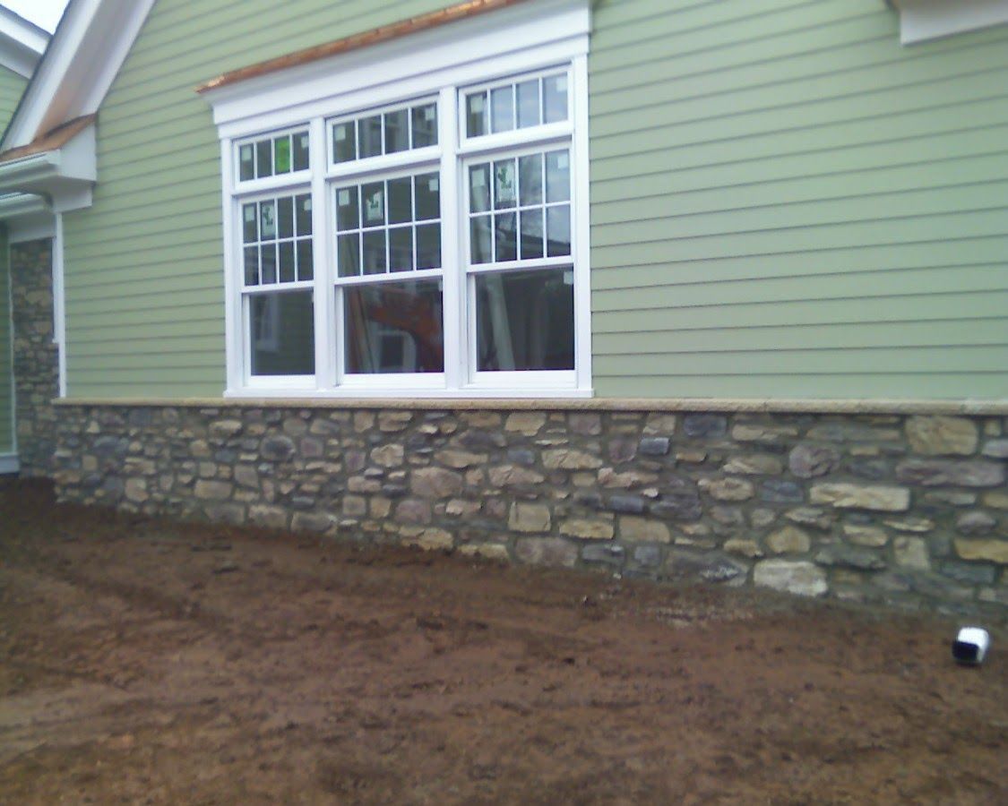 Stone foundation beneath green house siding and white-trimmed window over bare soil.
