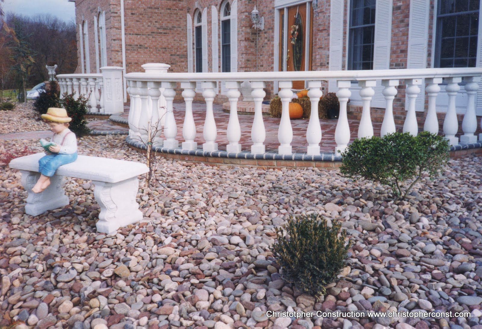Person sitting on a white stone bench in a rocky yard beside a brick house and white railing