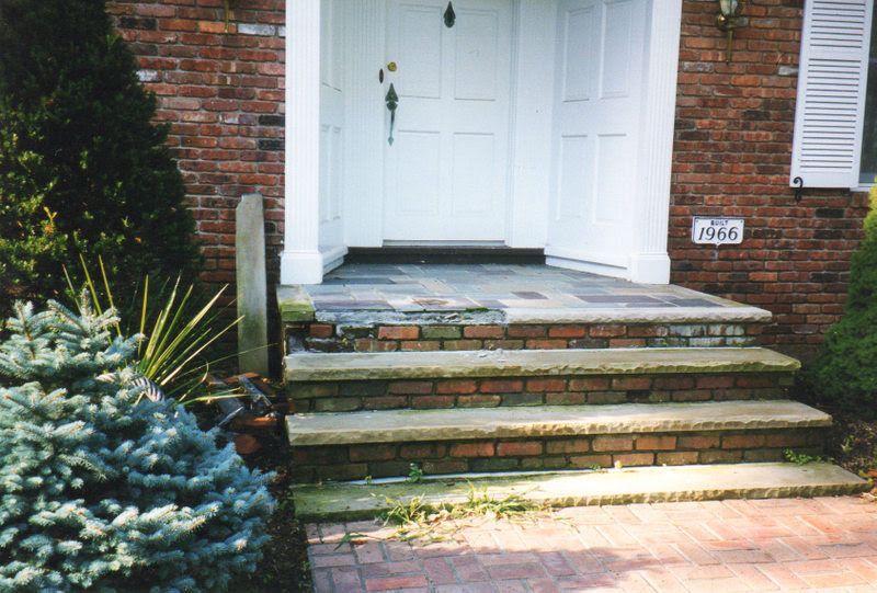 Front steps of a brick house leading to white double doors, with shrubs and a small walkway.