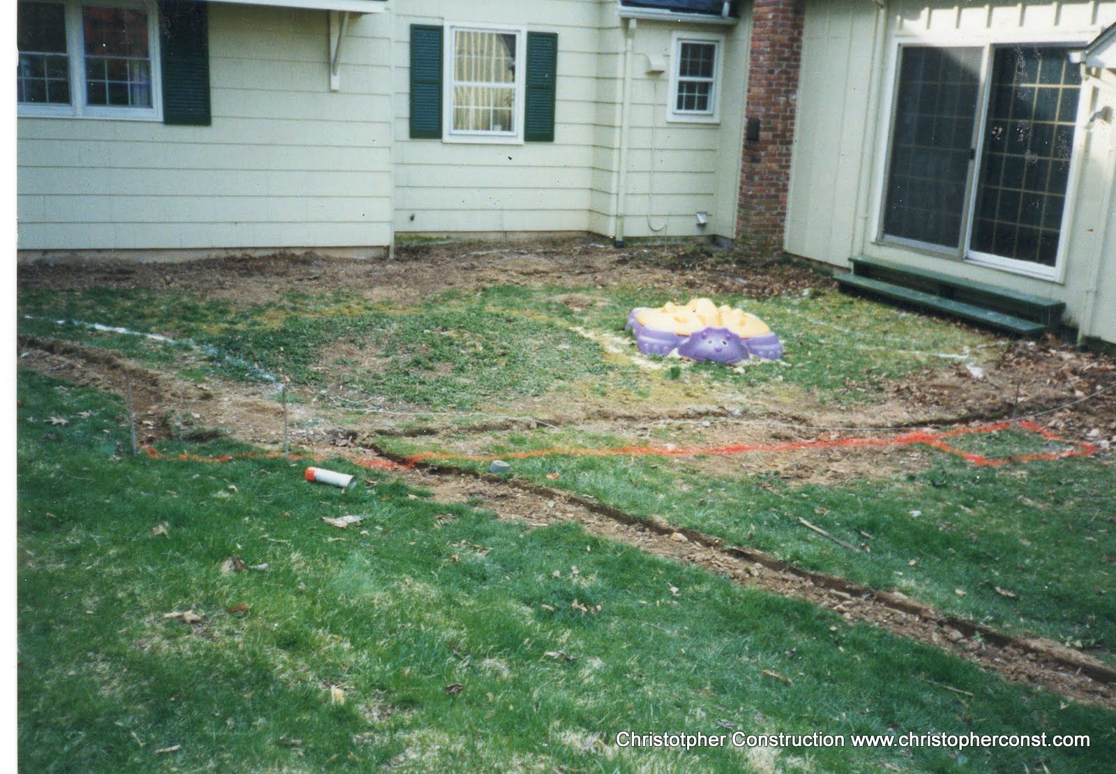 Backyard with patchy grass, a long hose or cable, and a purple object near a house and patio door.
