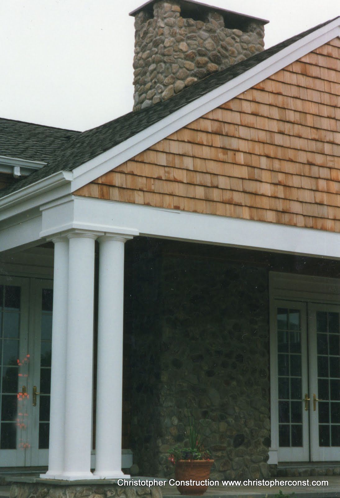Front porch of a house with white columns, cedar-shingle siding, and a stone chimney
