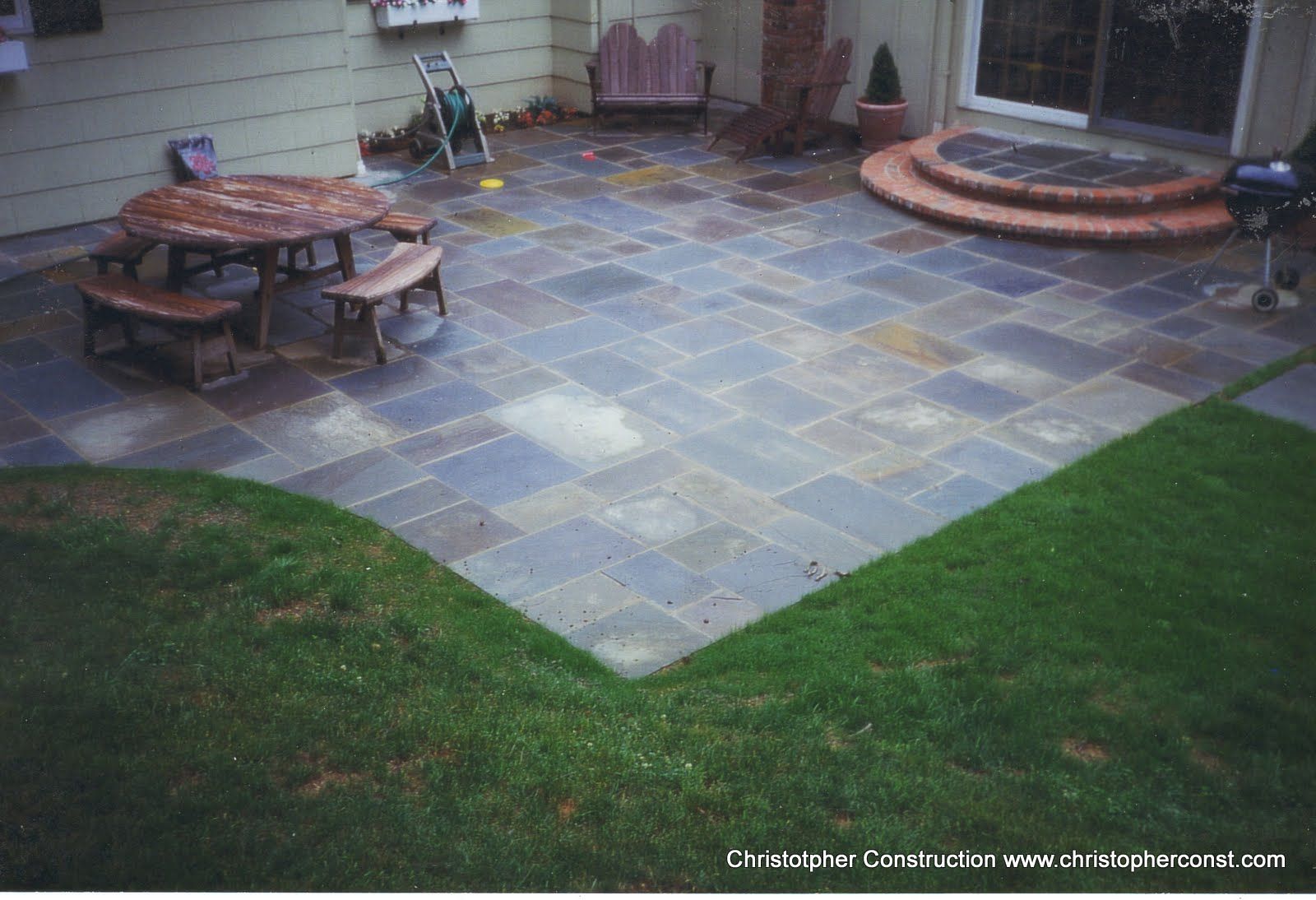 Backyard patio with slate pavers, circular fire pit seating, and brick steps by a house.