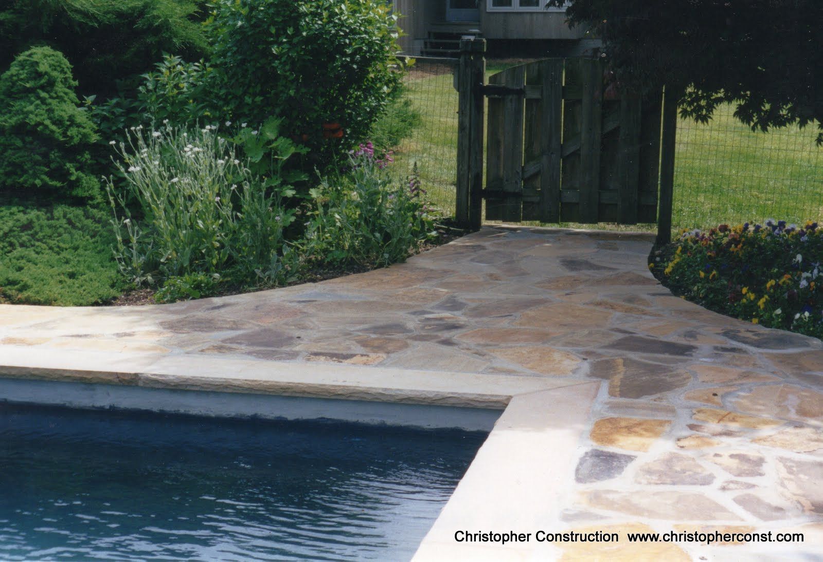 Backyard stone patio beside a swimming pool, with a wooden gate and lush greenery.