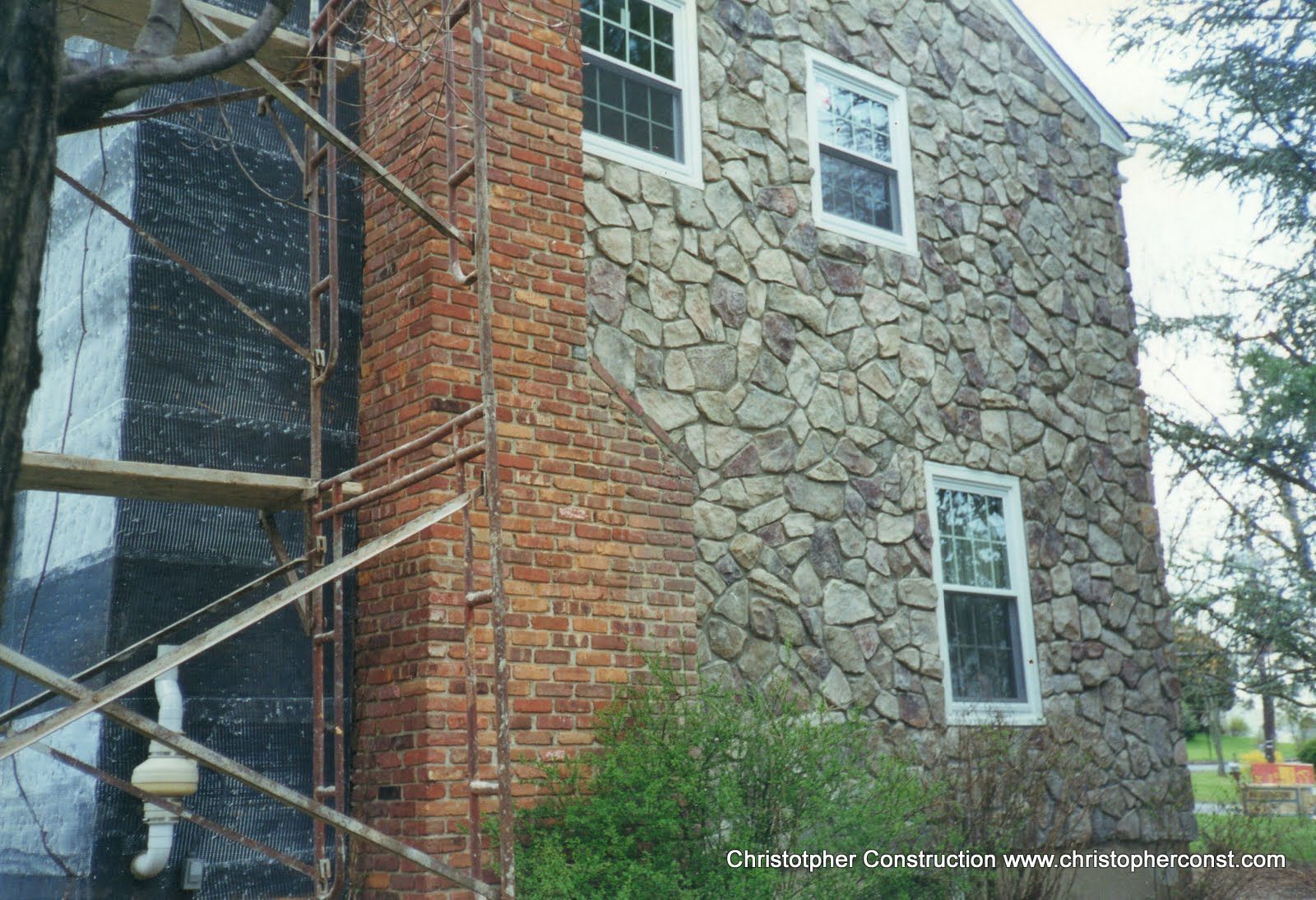 Stone-and-brick house exterior with windows, partly covered by scaffolding and netting