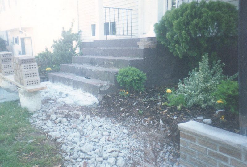 Front steps beside a house with a gravel walkway, shrubs, and a brick planter wall