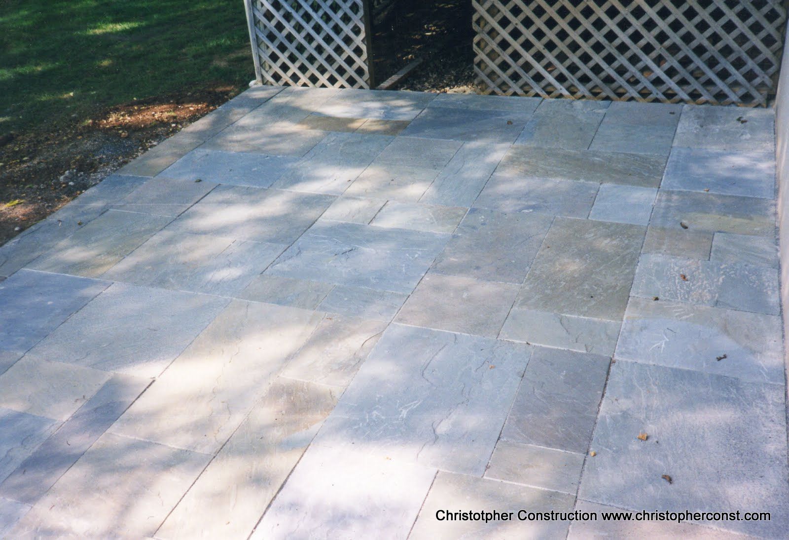 Stone patio with light and shadow, bordered by a lattice fence and surrounded by trees.
