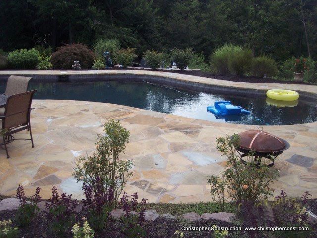 Backyard swimming pool with stone patio, lounge chair, and colorful float rings amidst greenery