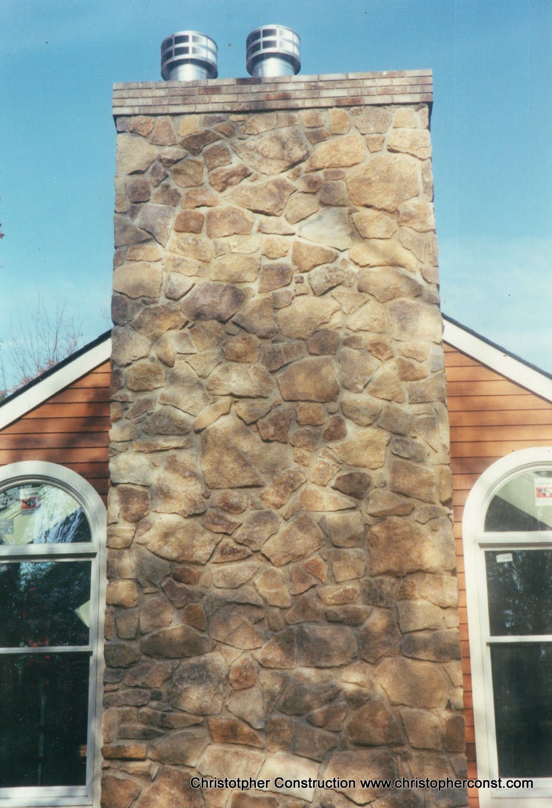 Stone chimney centered on a house roof, with two metal vents and arched windows on either side.