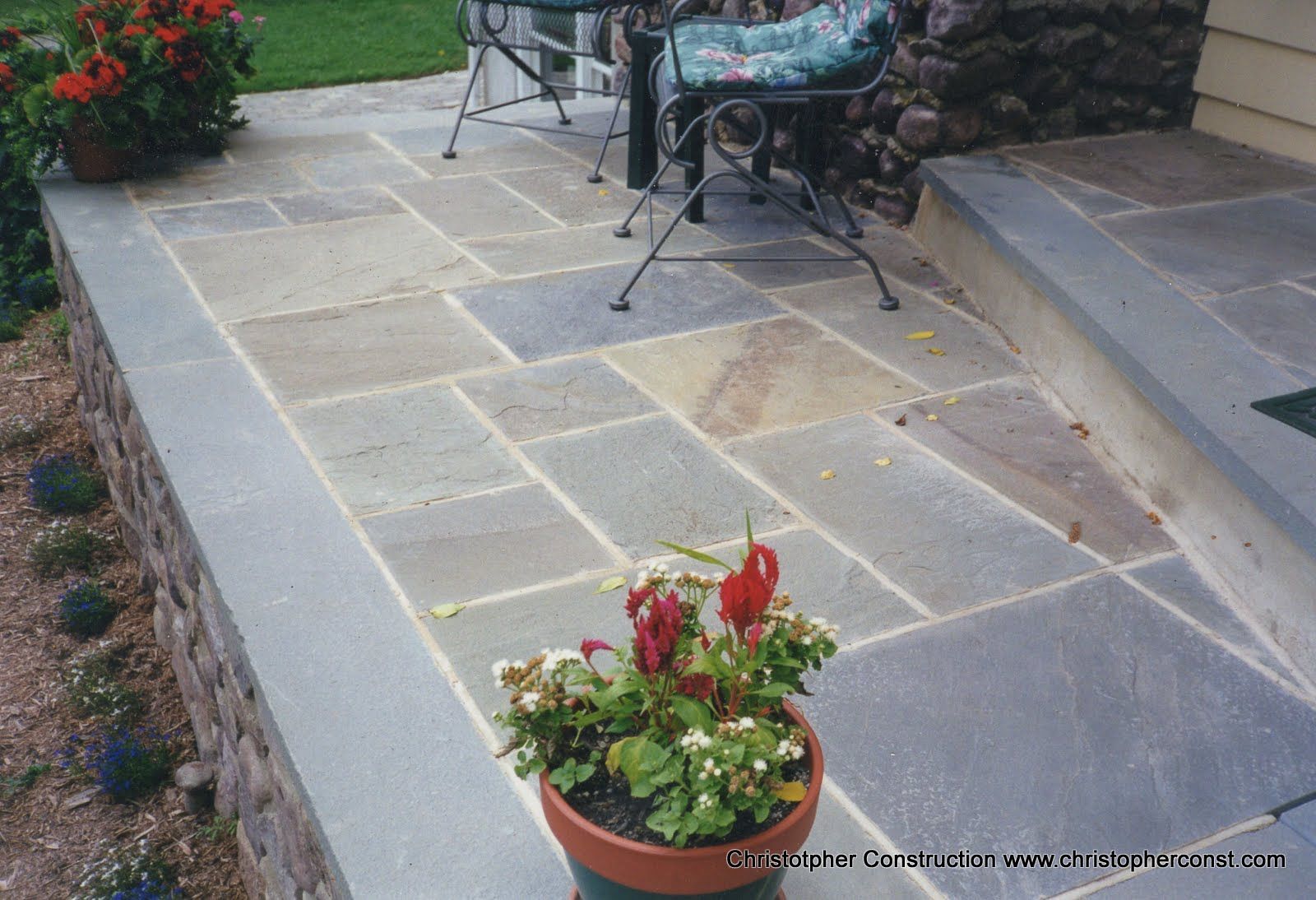 Stone patio with potted red flowers, outdoor chair, and garden steps at the side