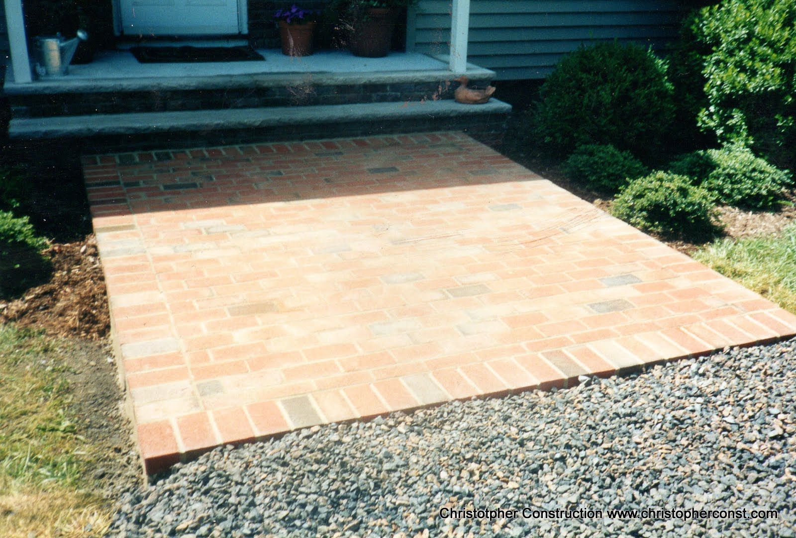 Brick patio walkway leading to a front porch, bordered by gravel and shrubs