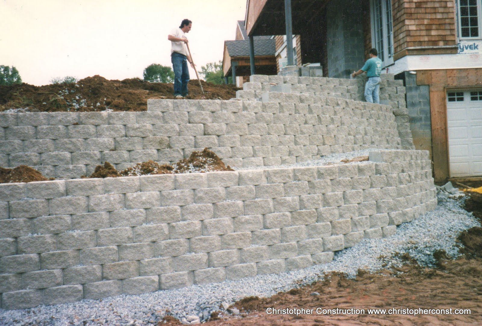 Person standing on a large terraced stone retaining wall beside a brick house under construction