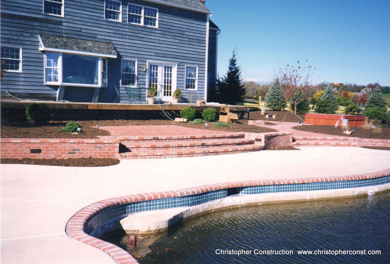 Backyard patio with curved pool beside a blue house and landscaped brick garden beds