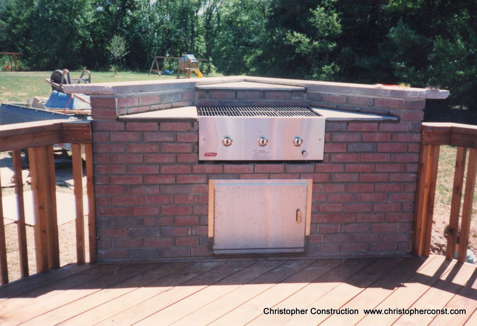 Brick outdoor grill with metal cabinet doors and side counters on a wooden deck, backed by trees
