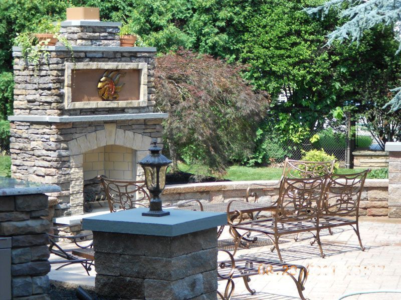 Stone patio with outdoor fireplace, black lantern, and wrought-iron chairs in a leafy garden