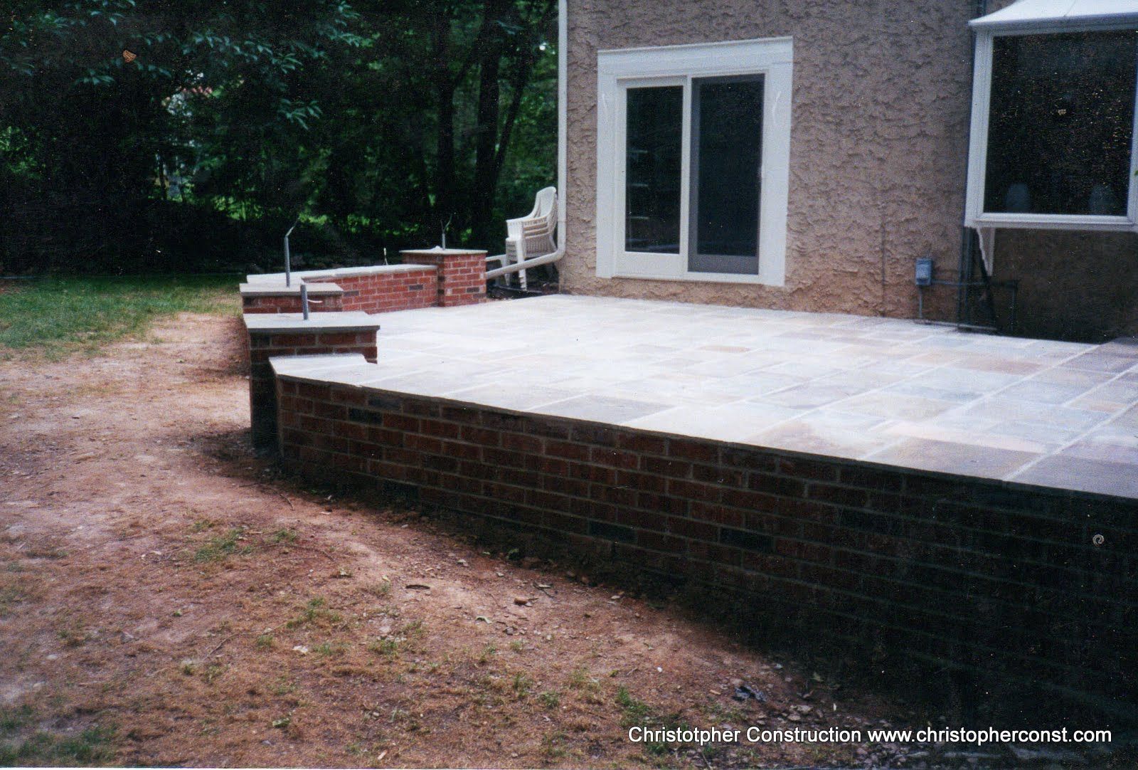 Brick patio with steps beside a house and wooded yard.