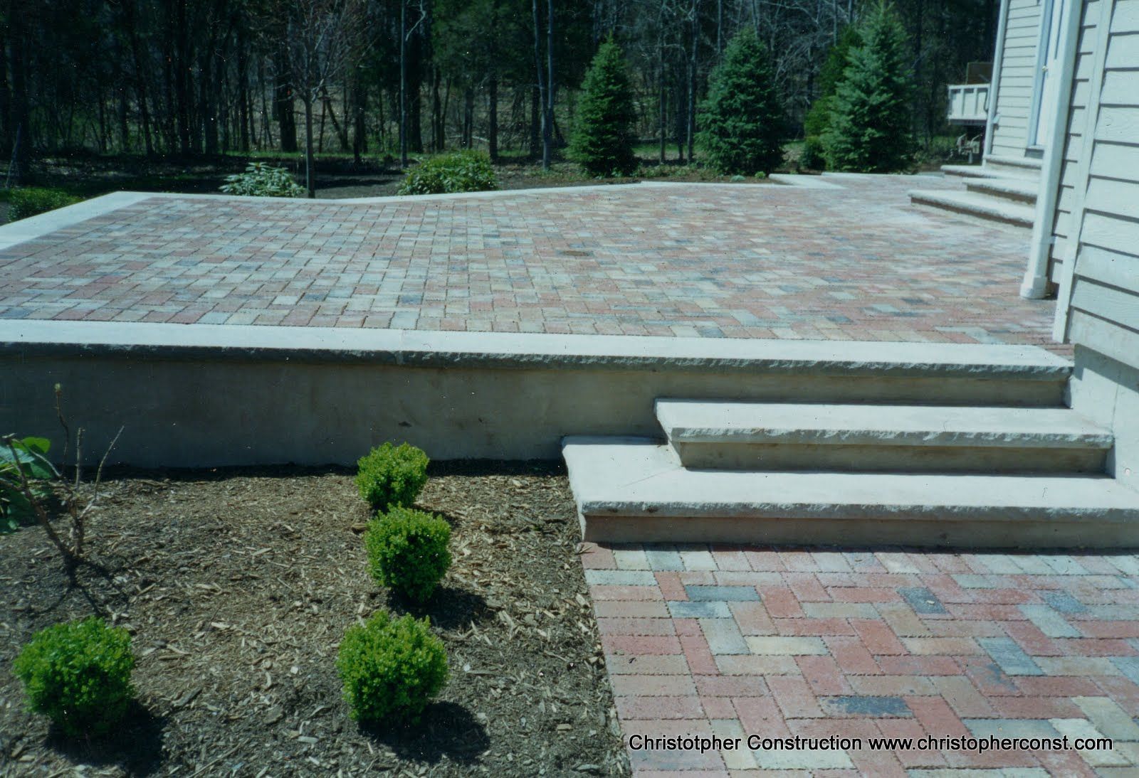 Patio with red brick pavers, concrete steps, and small shrubs beside a house