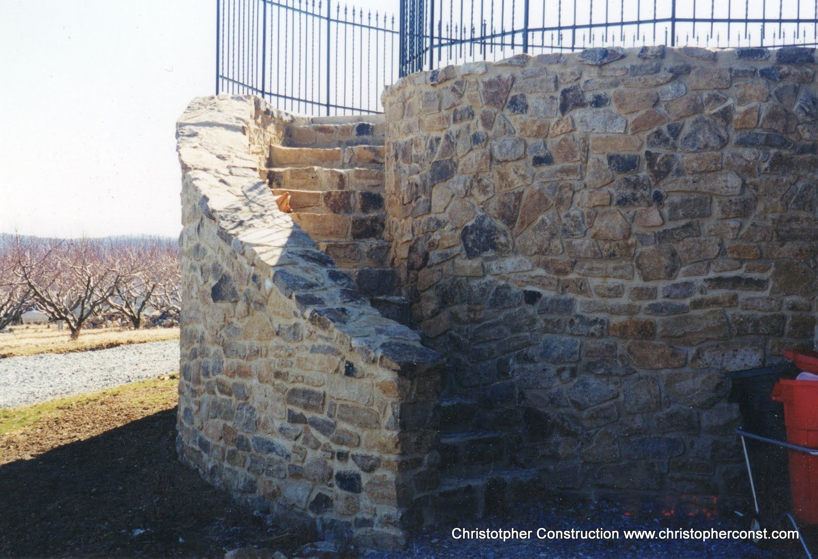 Stone retaining wall with a damaged corner beside a fence in a sunny outdoor area