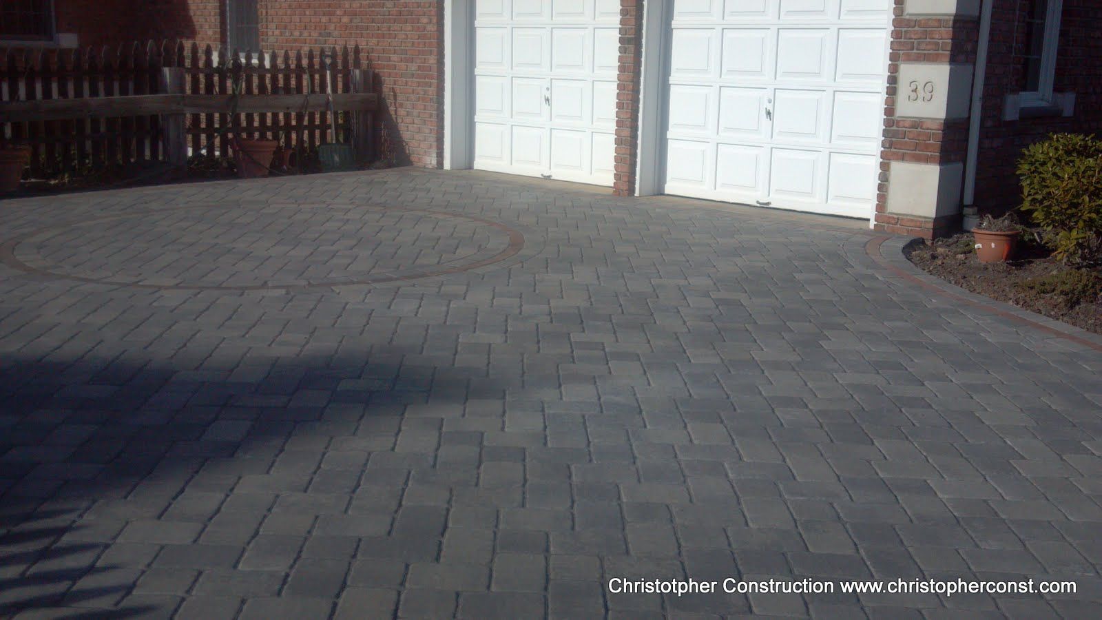Brick driveway leading to a house with white garage doors and a small garden bed on the right