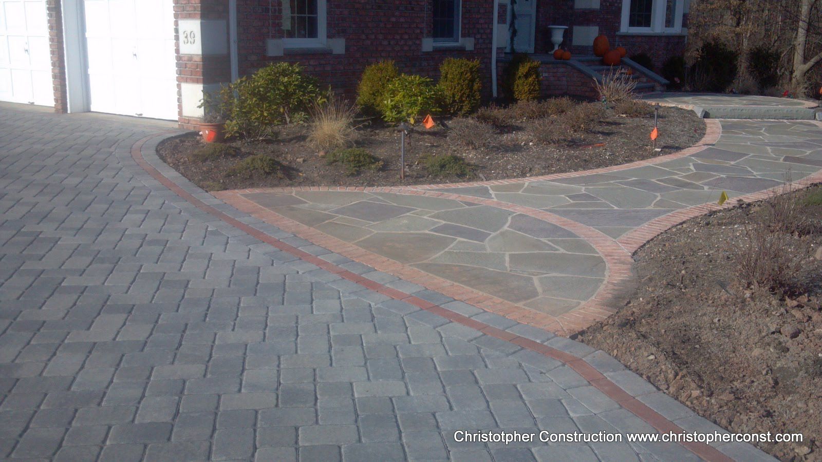 Curved brick walkway and stone patio beside a house with landscaped flower beds.