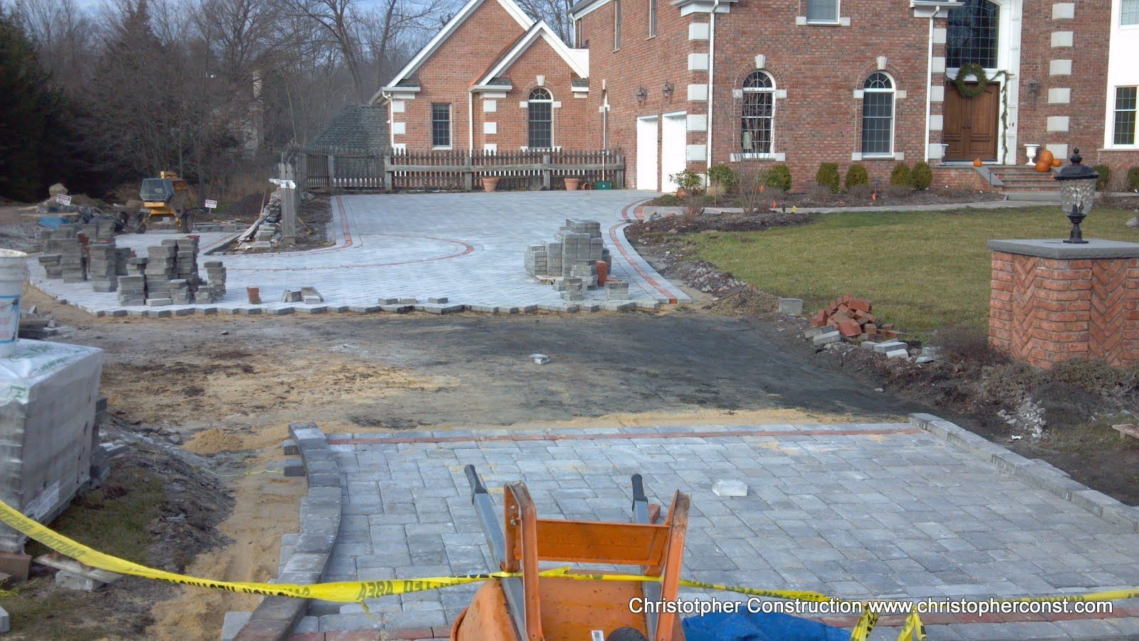 Partially paved walkway and driveway under construction in front of a brick building, with yellow caution tape.