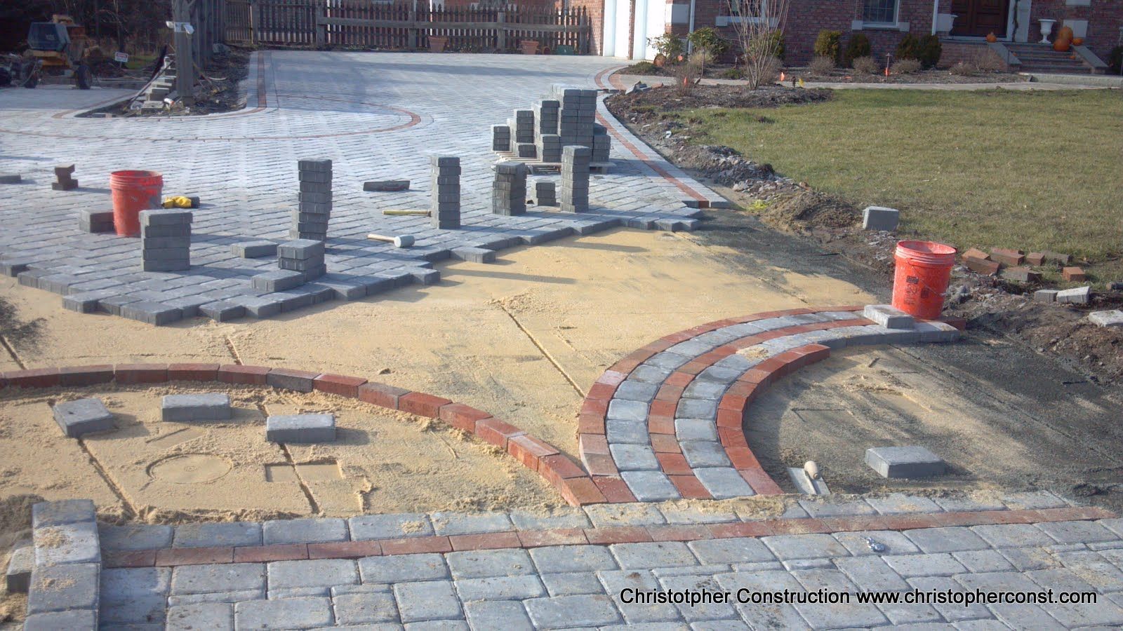 Curved brick walkway under construction with red buckets, gray posts, and a muddy paved area nearby