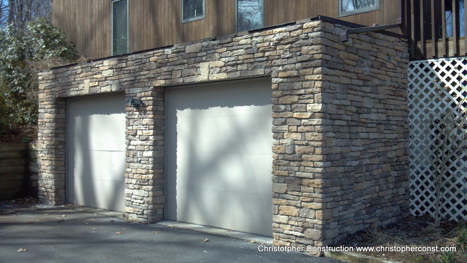 Stone garage with two white doors beside a driveway and lattice fence