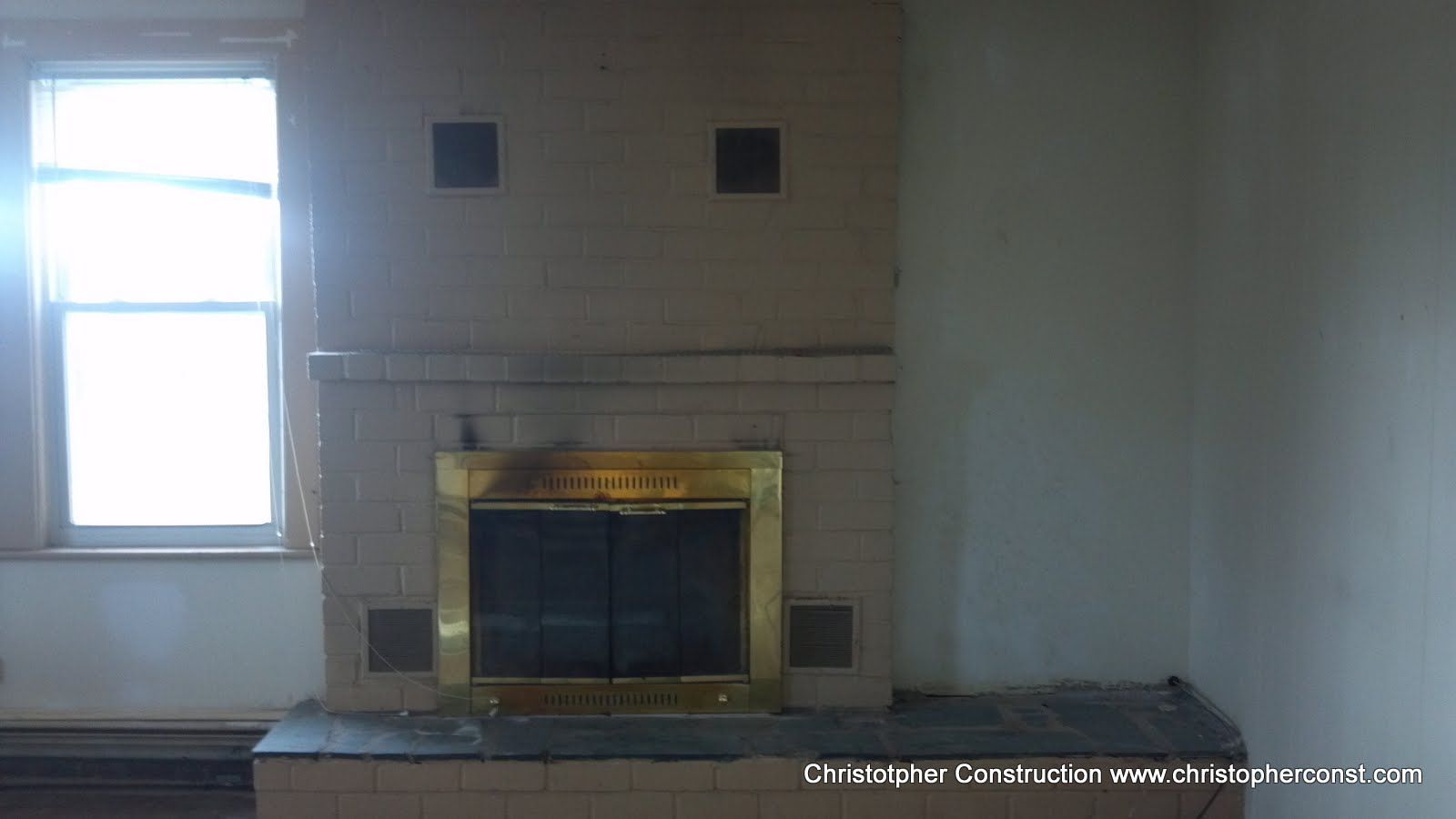 Empty room with a fireplace, two small wall vents, and a bright window on the left.