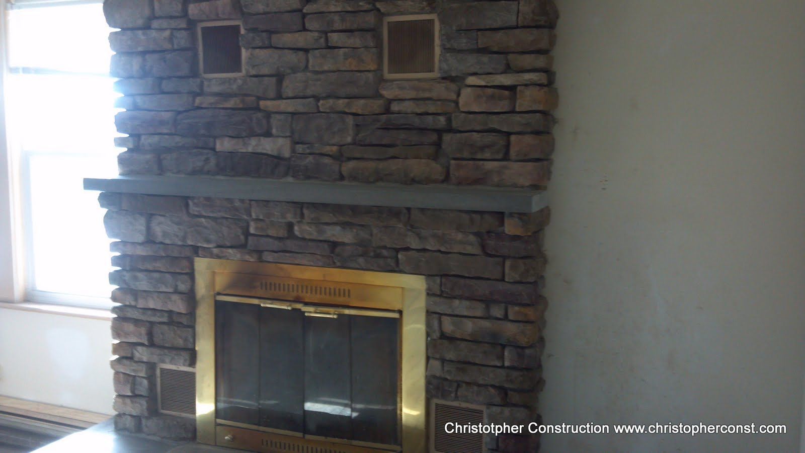 Stone fireplace with gold trim and glass doors in a room, with a window on the left and bare wall on the right