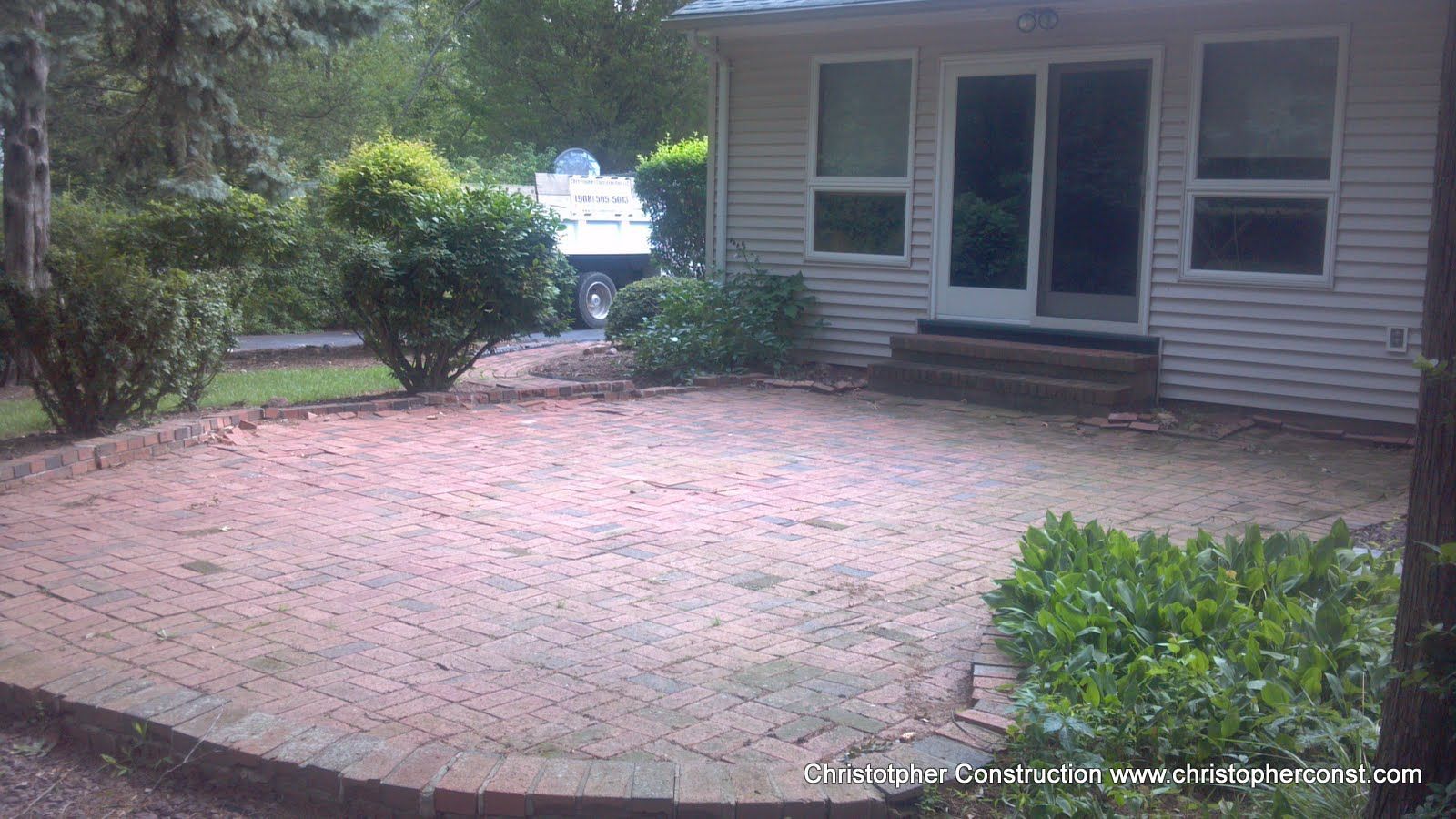 Patio with brick pavers beside a house, framed by shrubs and a bench in a leafy yard