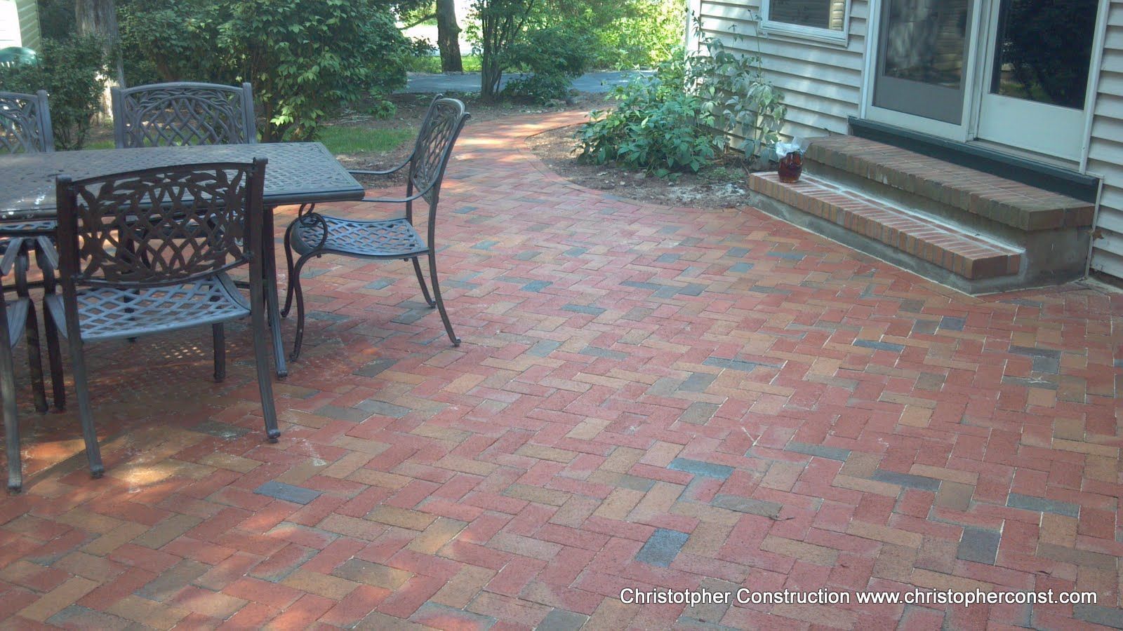 Brick patio beside a house with a black metal table and chairs, surrounded by greenery.