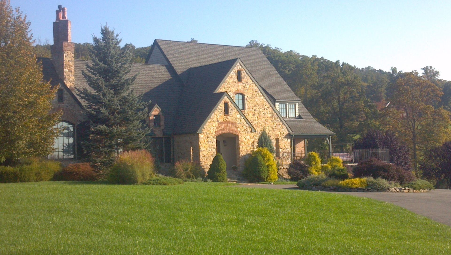 Stone house with a chimney and dormer windows beside a green lawn and trees in sunlight