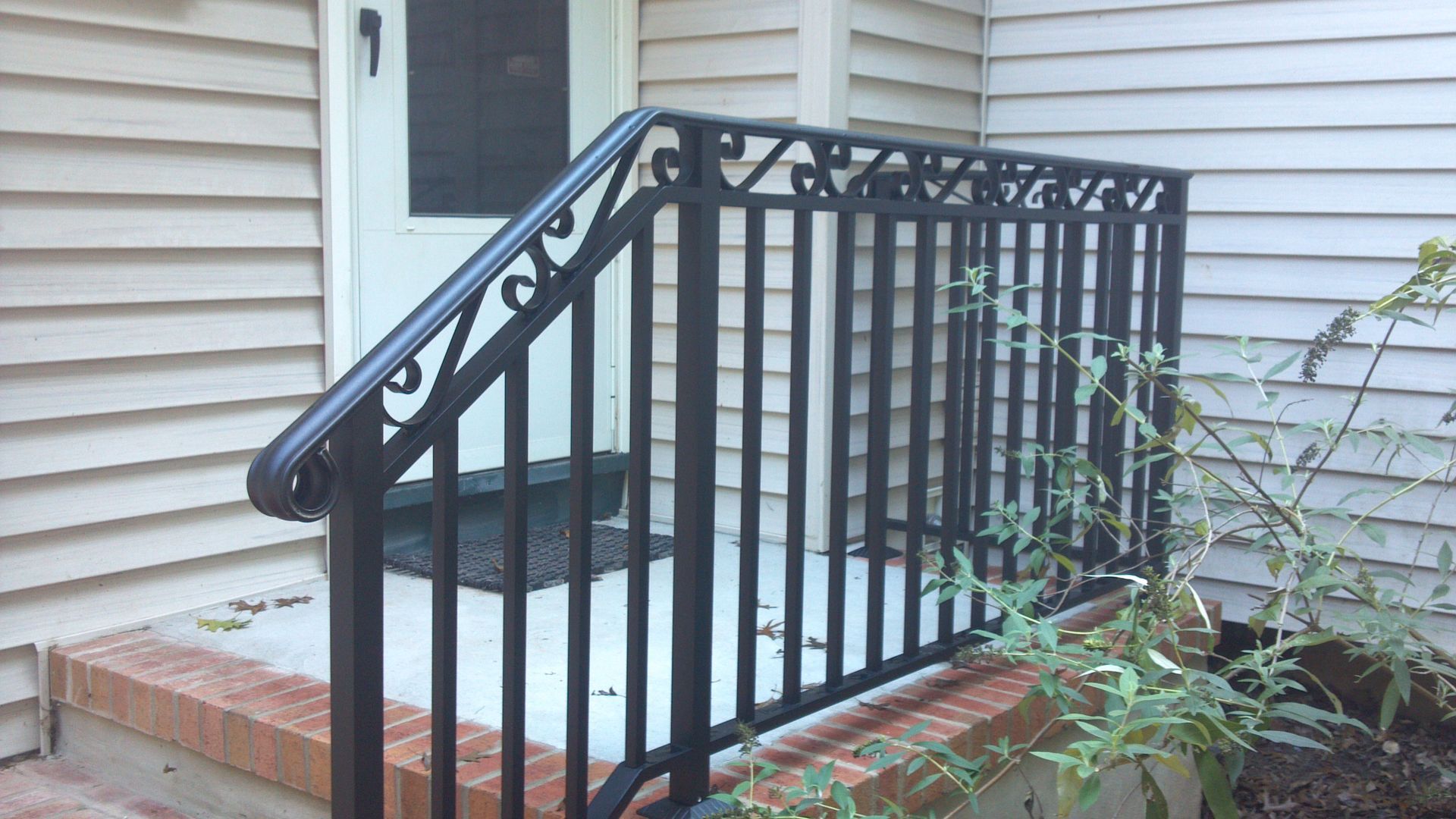 Black metal porch railing and steps on a white house with a brick base and small plants nearby