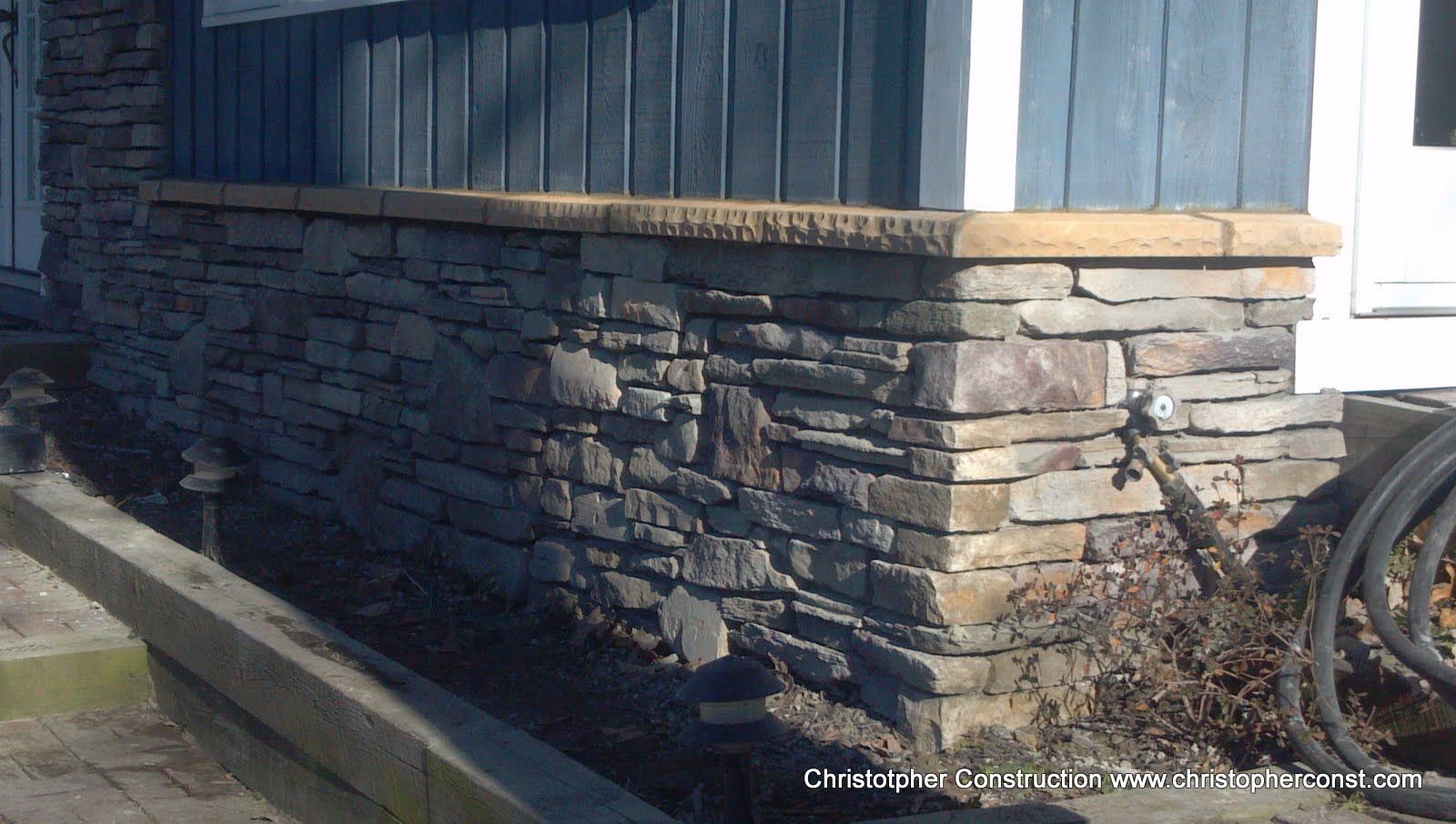 Stone-clad house exterior with blue siding, white trim, and a hose reel by the wall.
