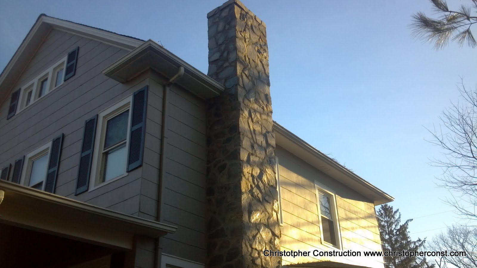 Two-story beige house with a tall stone chimney and blue shutters in daylight