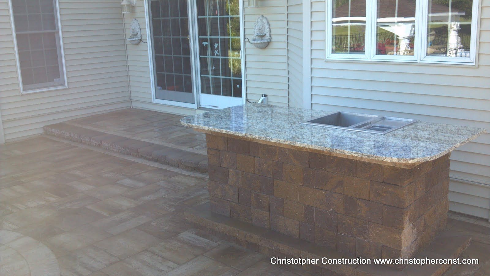 Back patio with stone pavers and a raised outdoor kitchen island beside sliding glass doors