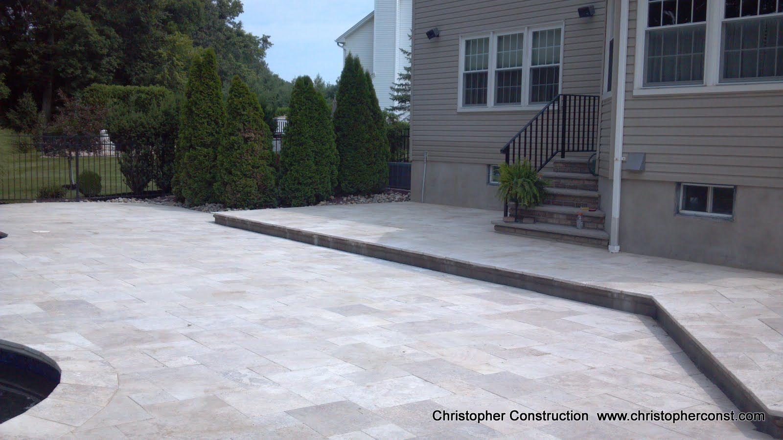 Concrete driveway beside a beige house, with a staircase and small side yard bordered by tall shrubs.