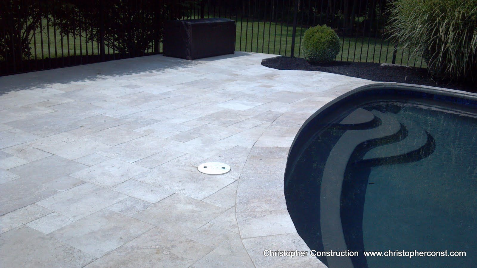Light stone patio beside a swimming pool with dark water and a drain cover in the center.
