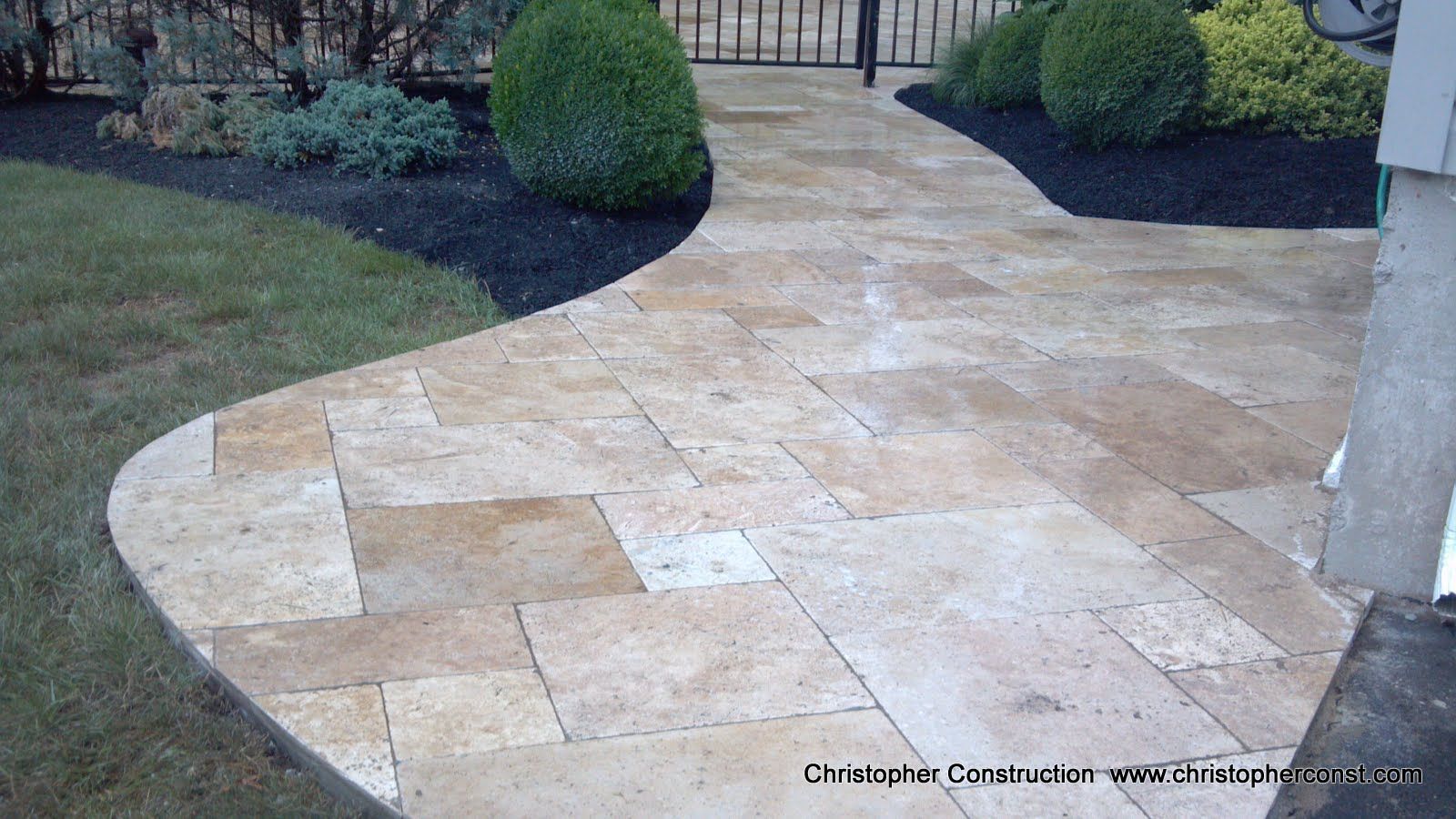 Curved beige stone walkway through a landscaped yard with shrubs and a fence