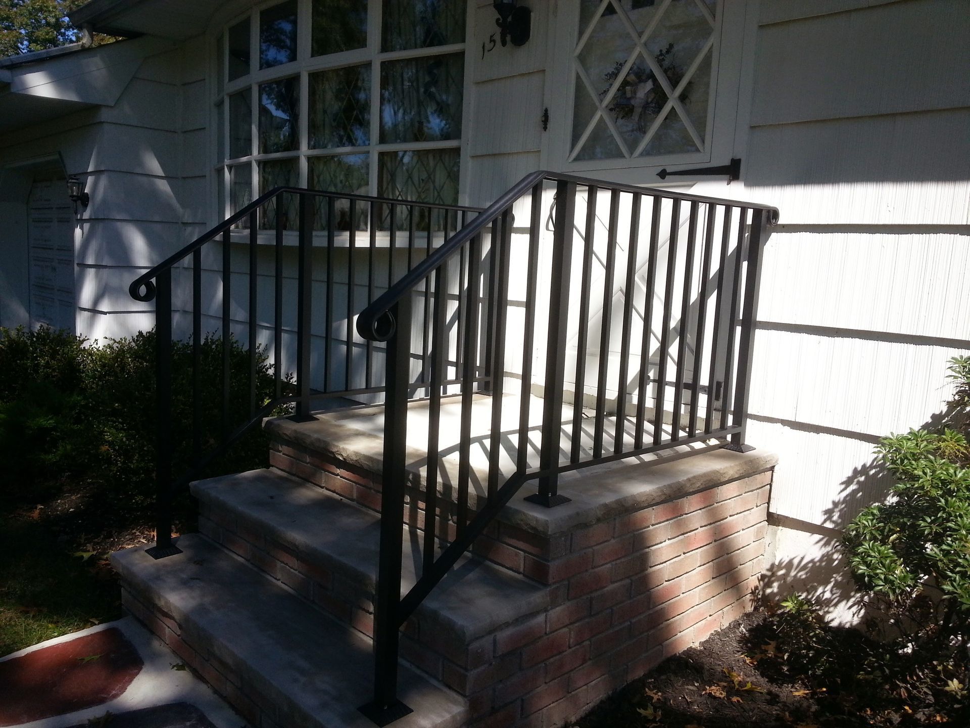 Brick porch steps with black metal railings beside a white house in sunlight