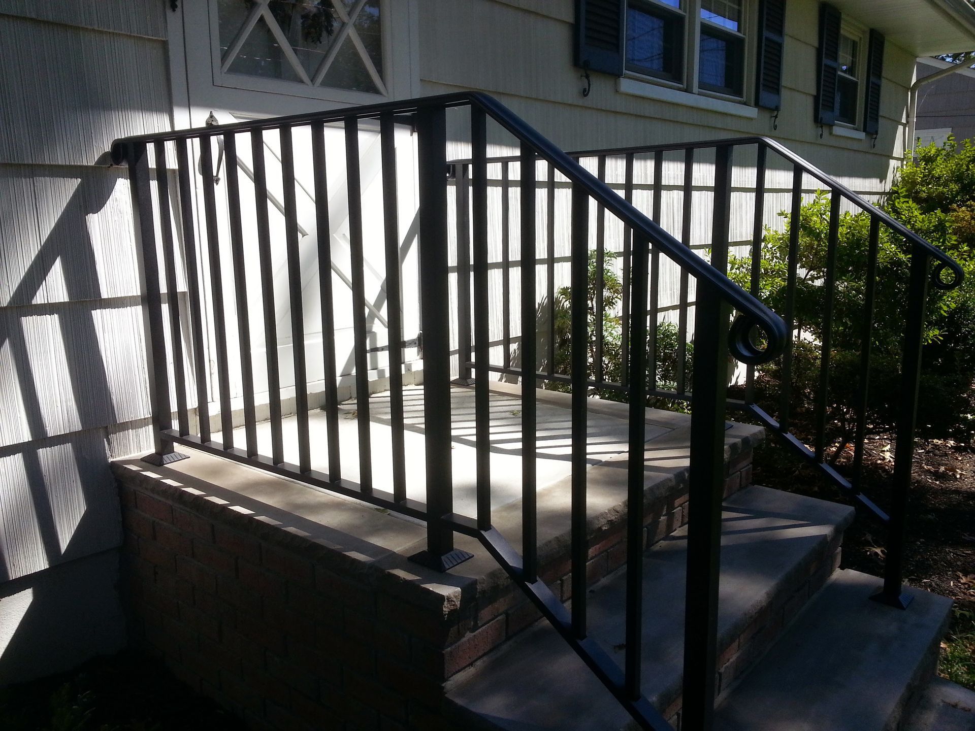 Black metal handrail and stairs leading to a raised porch beside a house in sunlight