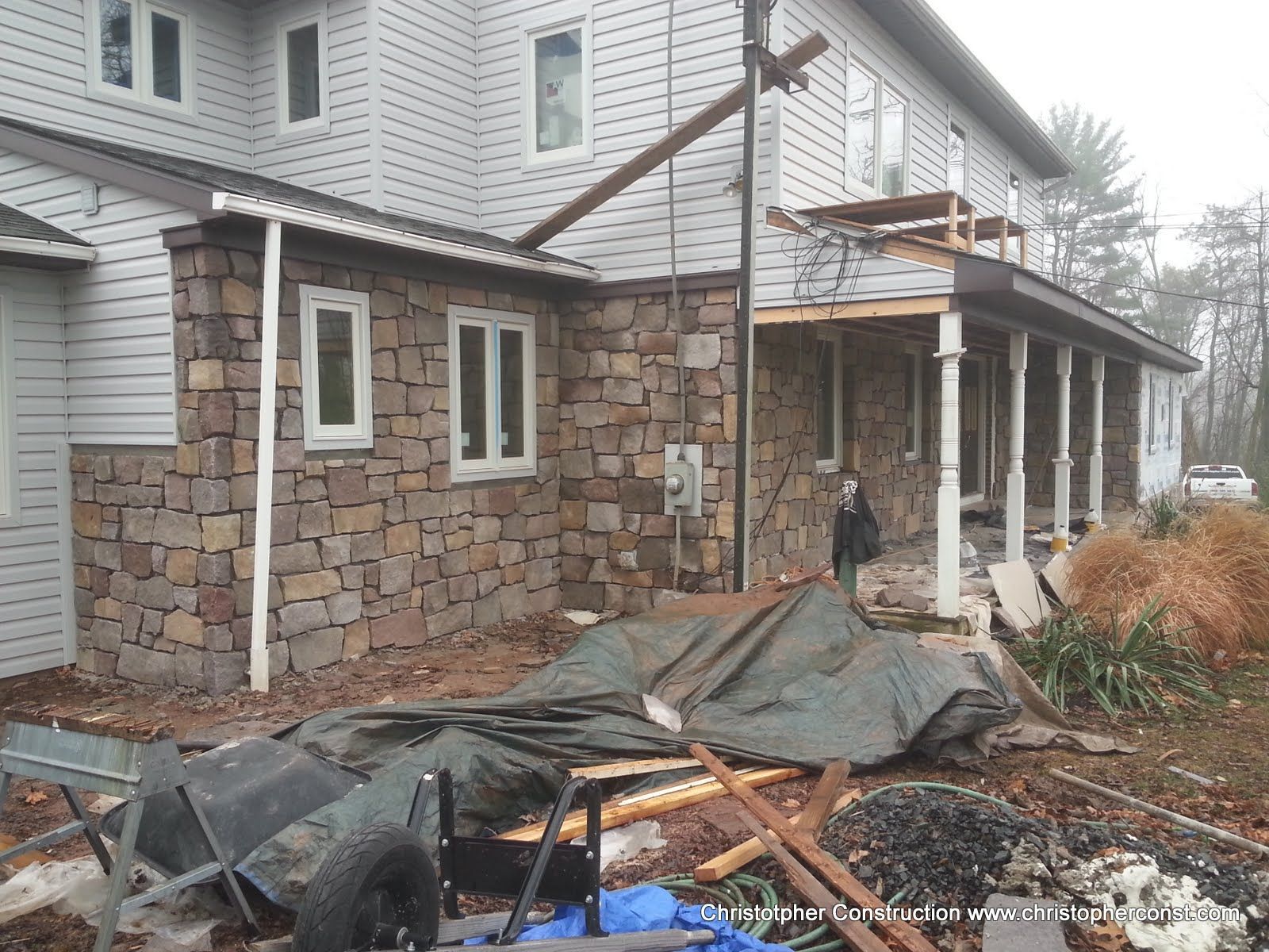 Stone house under construction with scaffolding, tarp-covered materials, and unfinished porch columns