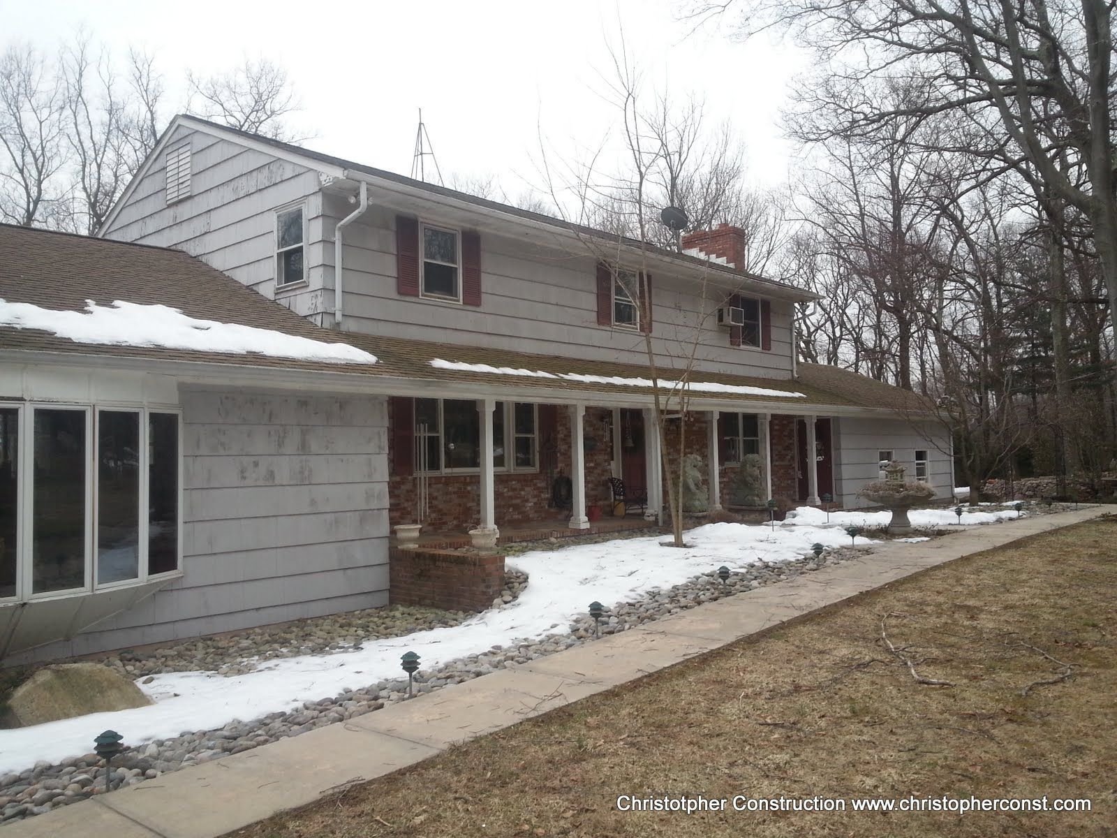 Two-story house with gray siding and brick porch in a snowy yard with bare trees