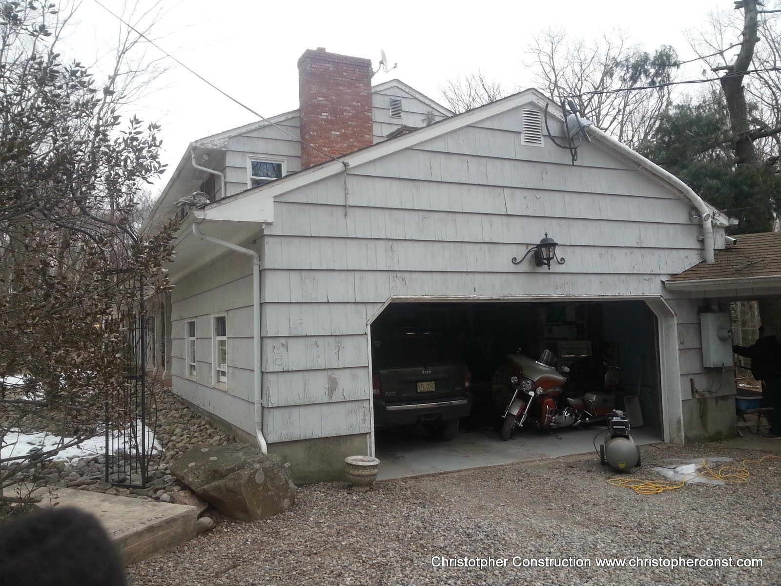 White garage with open door; black SUV and motorcycle inside, in a wooded residential driveway.