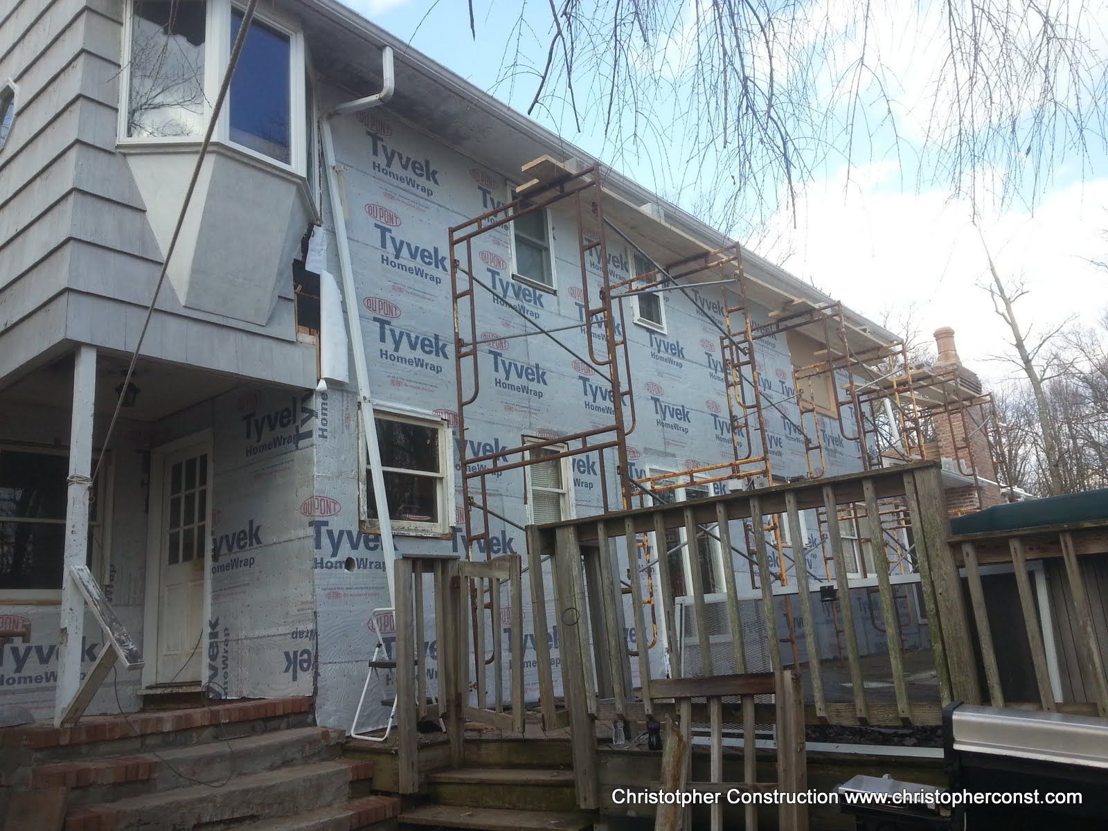 House under construction with exposed framing, wrap, and scaffolding beside a fenced yard