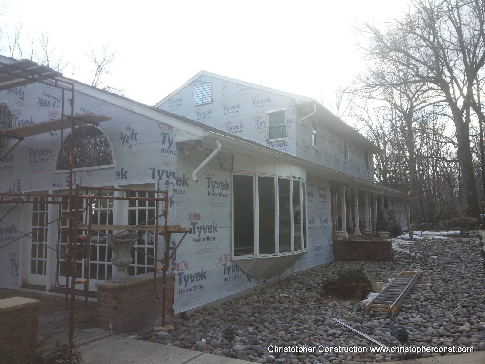 Partially built house wrapped in Tyvek, with porch columns and leaf-covered ground beside bare trees