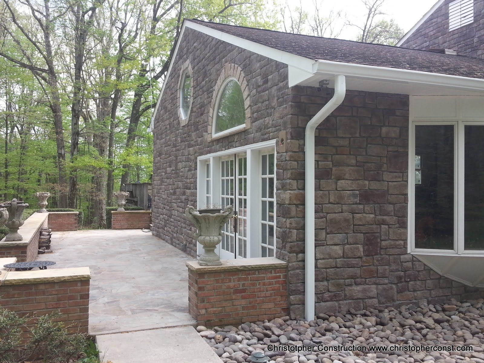 Stone house patio with white-trim windows and a walkway beside a wooded backyard