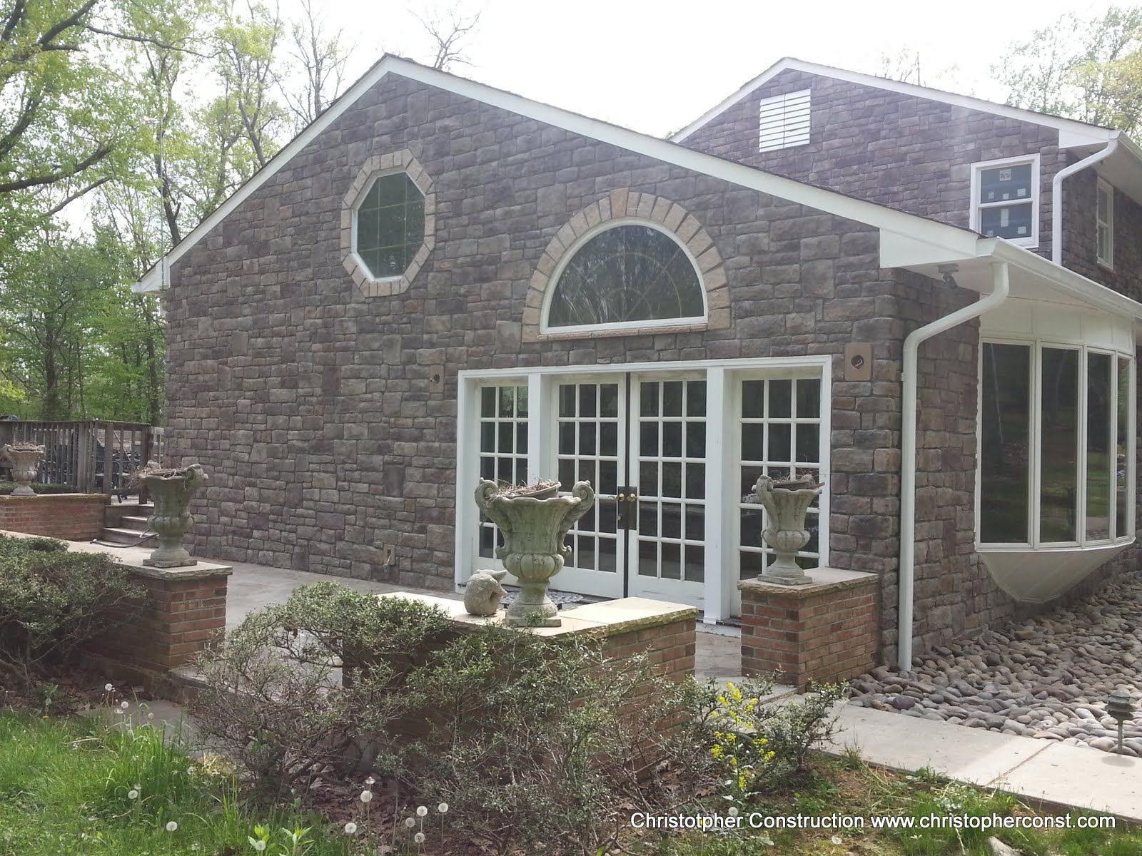 Stone house exterior with a front patio, French doors, and arched windows surrounded by trees
