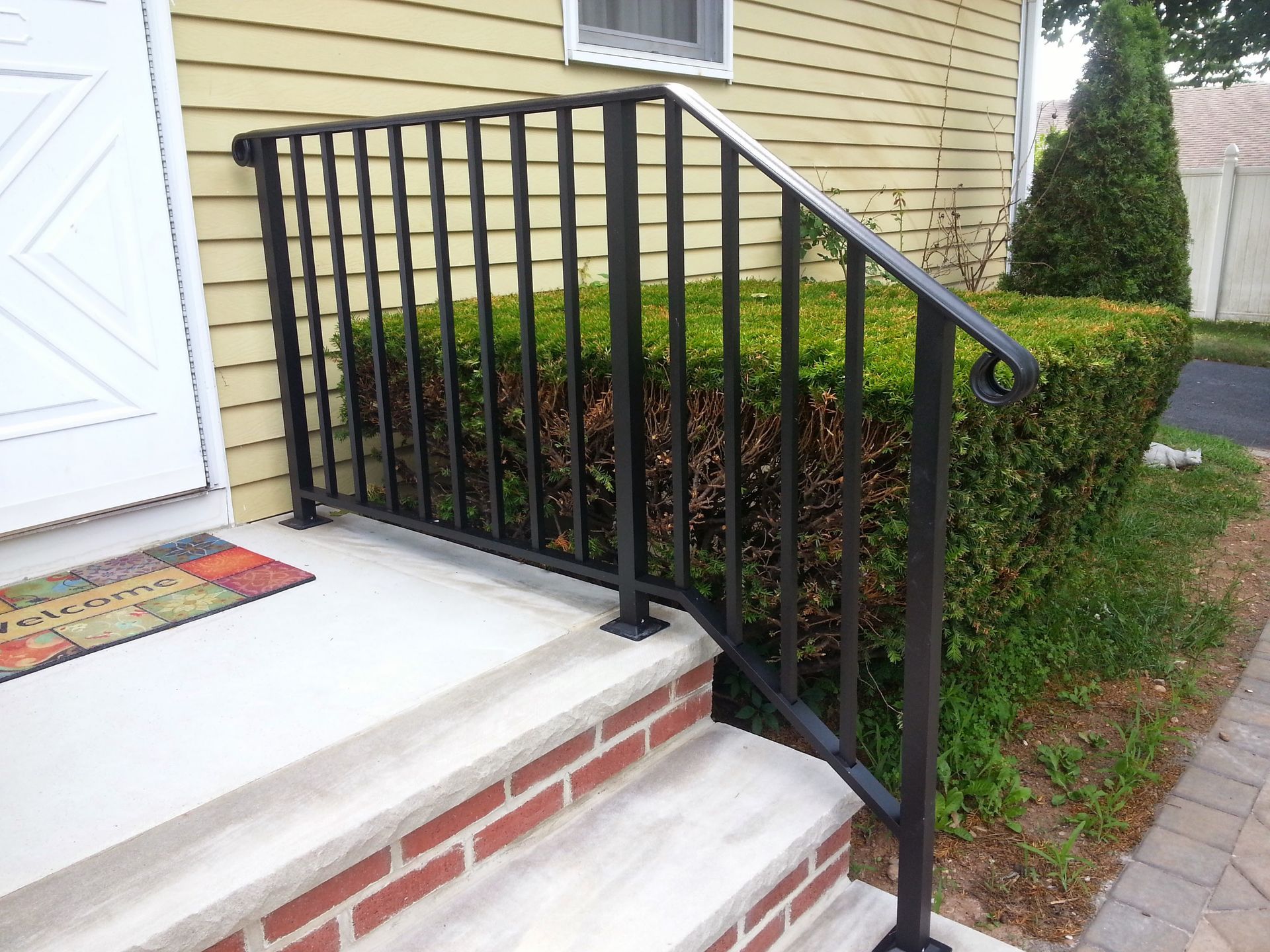Front steps with black metal railing beside a beige house and trimmed hedges